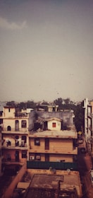 A residential area features multi-story buildings in an urban setting. Some buildings have balconies with decorative railings and rooftops adorned with antennas and satellite dishes. The scene is under a vast sky with a flock of birds flying in the distance. In the backdrop, distant trees and a hazy skyline are visible.