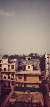 A residential area features multi-story buildings in an urban setting. Some buildings have balconies with decorative railings and rooftops adorned with antennas and satellite dishes. The scene is under a vast sky with a flock of birds flying in the distance. In the backdrop, distant trees and a hazy skyline are visible.