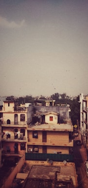 A residential area features multi-story buildings in an urban setting. Some buildings have balconies with decorative railings and rooftops adorned with antennas and satellite dishes. The scene is under a vast sky with a flock of birds flying in the distance. In the backdrop, distant trees and a hazy skyline are visible.