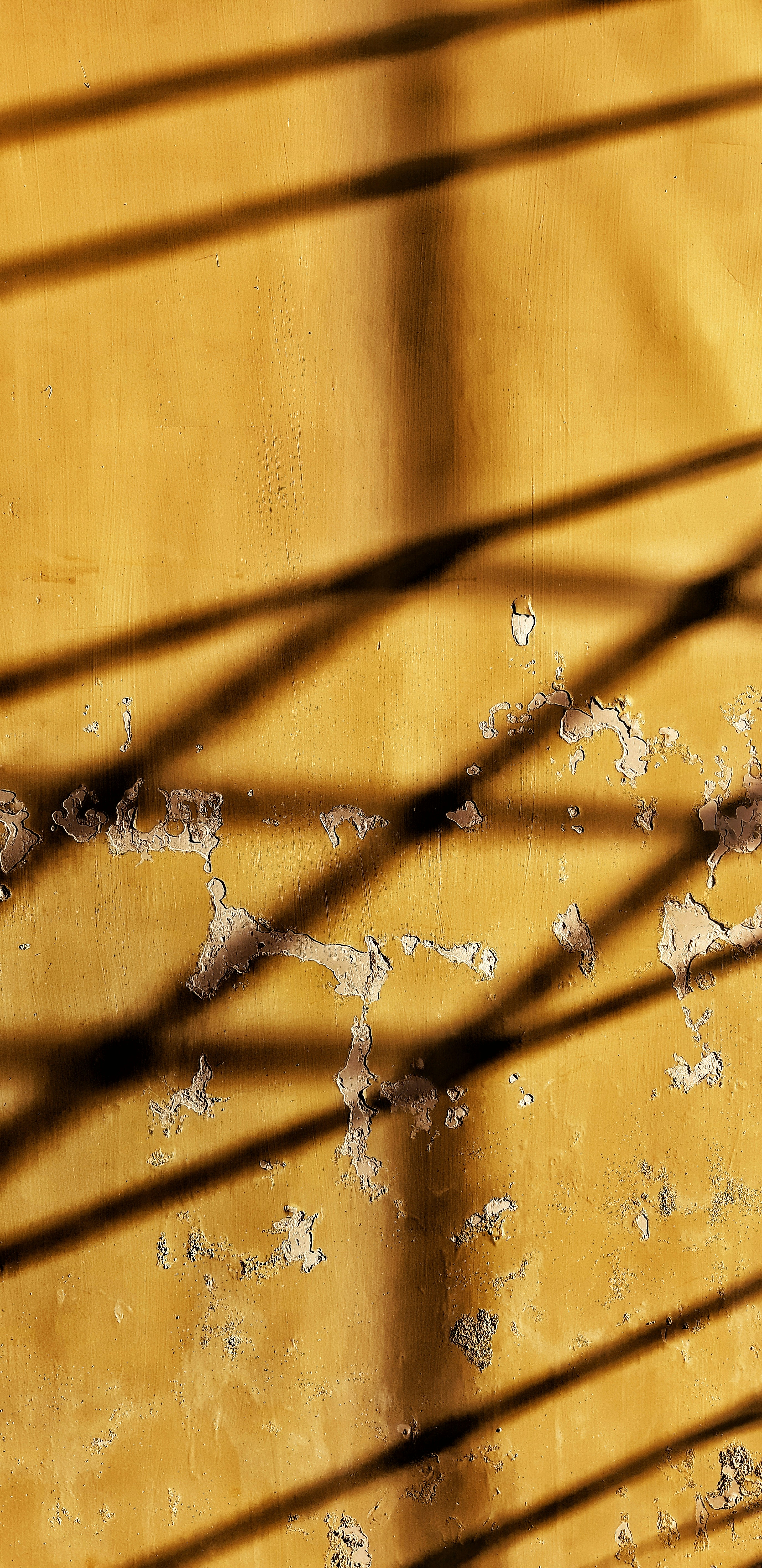 Weathered yellow wall with peeling patches and diagonal shadows creating abstract texture and rhythm. Color and texture emphasize aged surface with strong diagonal contrast.