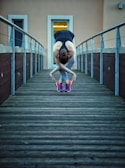 A person stretches in a forward bend on a wooden walkway. They wear athletic clothing including a black tank top and colorful sneakers. The railings and an entrance with a bright light are visible in the background.