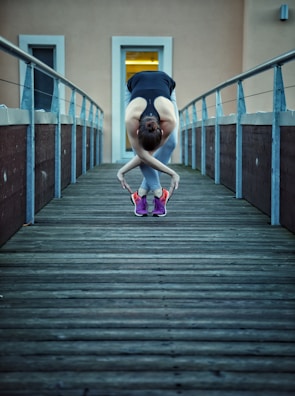 A person stretches in a forward bend on a wooden walkway. They wear athletic clothing including a black tank top and colorful sneakers. The railings and an entrance with a bright light are visible in the background.