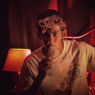 A person is exhaling smoke in a dimly lit room, creating a mysterious and moody atmosphere. The lighting is warm, coming from a nearby lamp, and casts red and orange hues on the surroundings. The person is wearing a light-colored t-shirt.