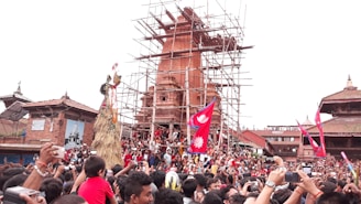 A large crowd gathered in front of a historic structure under restoration, with scaffolding visible. People are celebrating, waving red flags with a white emblem, likely during a festival. Traditional architecture is surrounding the scene, and individuals are capturing the event with their phones.