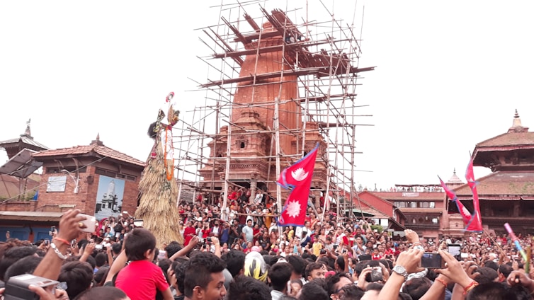 A large crowd gathered in front of a historic structure under restoration, with scaffolding visible. People are celebrating, waving red flags with a white emblem, likely during a festival. Traditional architecture is surrounding the scene, and individuals are capturing the event with their phones.
