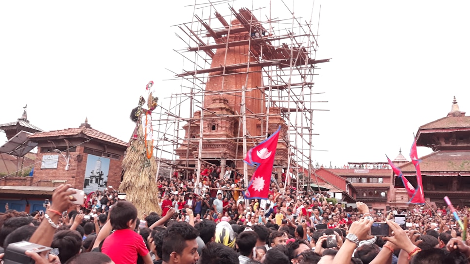 A large crowd gathered in front of a historic structure under restoration, with scaffolding visible. People are celebrating, waving red flags with a white emblem, likely during a festival. Traditional architecture is surrounding the scene, and individuals are capturing the event with their phones.