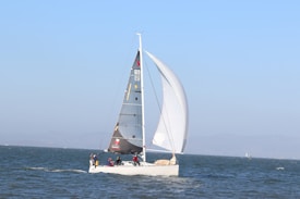 A sailboat glides on a vast body of water under clear blue skies. The boat is equipped with a tall mast and black sails with distinct markings. Several people are visible on the deck, actively managing the boat. The background reveals a faint shoreline with some hills or mountains.