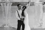A couple is dancing in a graceful pose. The woman is wearing a long white dress and a floral crown, and the man is wearing a white shirt with suspenders and dark pants. The setting is a textured, worn wall, adding an artistic backdrop to the scene.
