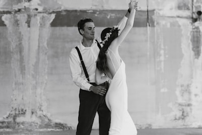 A couple is dancing in a graceful pose. The woman is wearing a long white dress and a floral crown, and the man is wearing a white shirt with suspenders and dark pants. The setting is a textured, worn wall, adding an artistic backdrop to the scene.