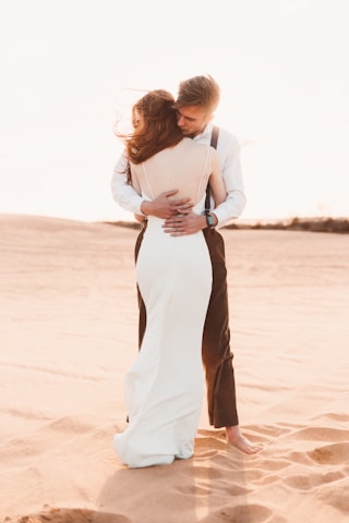 A couple embraces in a desert setting, with the woman wearing a white dress and the man dressed in a white shirt and suspenders. The warm sunlight creates a soft and romantic atmosphere as sand dunes stretch out in the background.