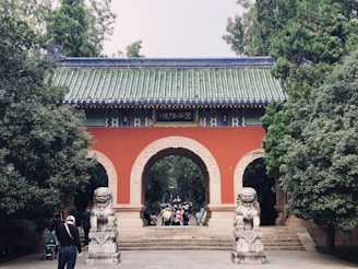 An ornate traditional Chinese gate with a green tiled roof and red walls arches over a pathway. Stone guardian lion statues flank the entrance, and several people are walking towards and through the gate. The scene is surrounded by lush green trees.