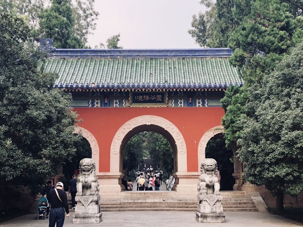 An ornate traditional Chinese gate with a green tiled roof and red walls arches over a pathway. Stone guardian lion statues flank the entrance, and several people are walking towards and through the gate. The scene is surrounded by lush green trees.