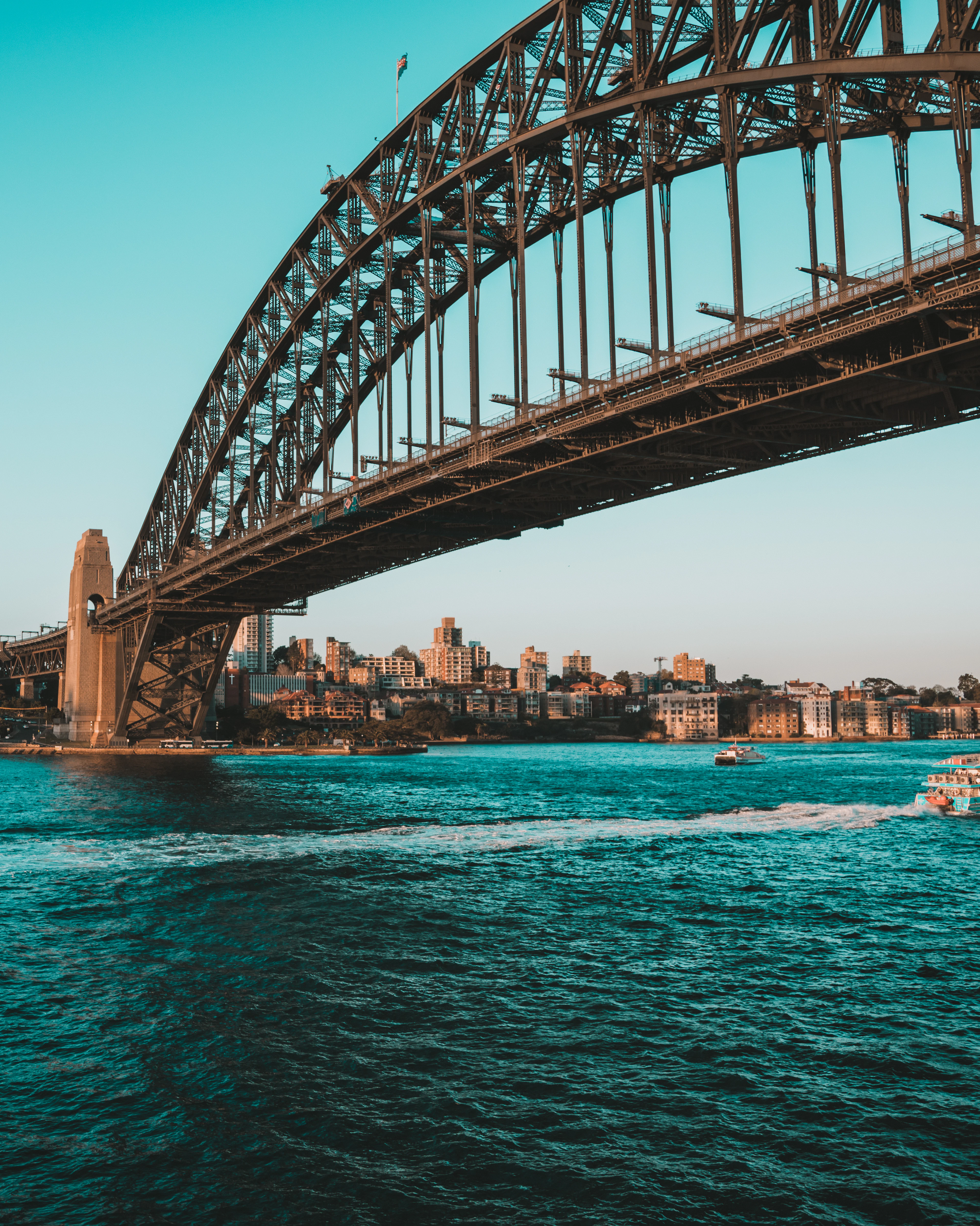 The steel arch of the Sydney Harbour Bridge seen from close to the water, with the harbourside shoreline and turquoise water beneath it