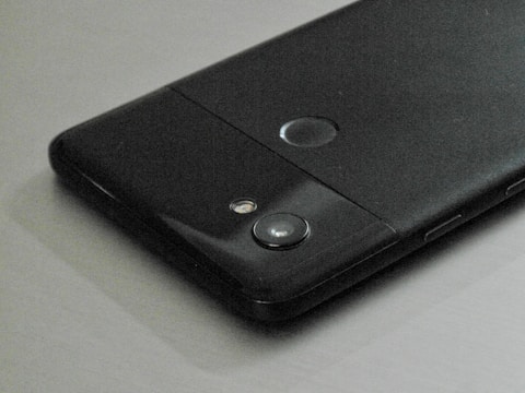 A close-up view of a black smartphone placed on a flat surface, showing its rear side with a camera lens, flash, and a circular fingerprint sensor.