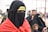 A group of people are dressed in traditional attire, with the foreground focused on a person wearing a black veil covering the face except for the eyes, and a red garment draped over the shoulders. A yellow headband with Arabic script is visible. In the background, more people are present, including a child, all wearing similar dark clothing.