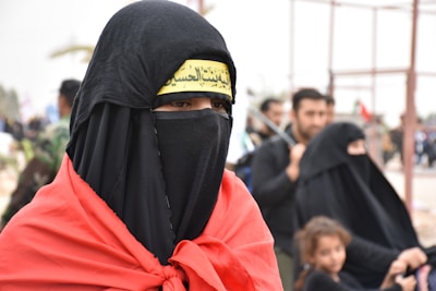 A group of people are dressed in traditional attire, with the foreground focused on a person wearing a black veil covering the face except for the eyes, and a red garment draped over the shoulders. A yellow headband with Arabic script is visible. In the background, more people are present, including a child, all wearing similar dark clothing.
