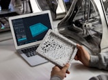 A person holds a detailed, intricately designed 3D-printed object, with a laptop displaying a 3D modeling software. The background features a 3D printer, suggesting a design and production process.