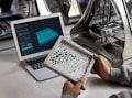 A person holds a detailed, intricately designed 3D-printed object, with a laptop displaying a 3D modeling software. The background features a 3D printer, suggesting a design and production process.