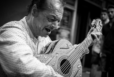 A man passionately playing an intricately decorated guitar in an outdoor setting. He appears to be deeply immersed in the music, with his eyes closed and mouth open, possibly singing. The scene captures a candid moment of emotional expression.