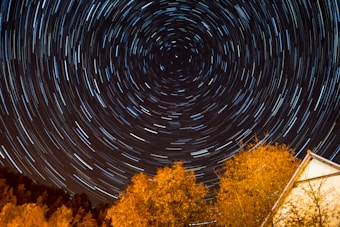 A mesmerizing display of star trails forming circular patterns in the night sky, centered around the North Star. Below, the silhouette of trees and a building with an orange glow from artificial light contrasts against the dark sky.