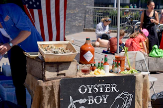 A street vendor stall with a rustic wooden crate holding a box of oysters on ice. Numerous bottles of hot sauce and a large pitcher are displayed next to a mixed arrangement of lemon wedges, cocktail sauce, and celery. An American flag hangs in the background, and several people are seen sitting nearby, some using their phones.