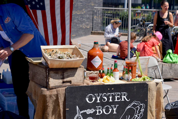 A street vendor stall with a rustic wooden crate holding a box of oysters on ice. Numerous bottles of hot sauce and a large pitcher are displayed next to a mixed arrangement of lemon wedges, cocktail sauce, and celery. An American flag hangs in the background, and several people are seen sitting nearby, some using their phones.