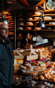 A busy cheese shop displaying a variety of cheeses stacked and arranged on a counter. A person is seen working behind the counter, and another individual is in the foreground. The shop is lit by hanging lamps, and shelves filled with wheels of cheese line the walls.