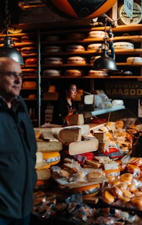 A busy cheese shop displaying a variety of cheeses stacked and arranged on a counter. A person is seen working behind the counter, and another individual is in the foreground. The shop is lit by hanging lamps, and shelves filled with wheels of cheese line the walls.