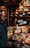A busy cheese shop displaying a variety of cheeses stacked and arranged on a counter. A person is seen working behind the counter, and another individual is in the foreground. The shop is lit by hanging lamps, and shelves filled with wheels of cheese line the walls.