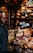 A busy cheese shop displaying a variety of cheeses stacked and arranged on a counter. A person is seen working behind the counter, and another individual is in the foreground. The shop is lit by hanging lamps, and shelves filled with wheels of cheese line the walls.