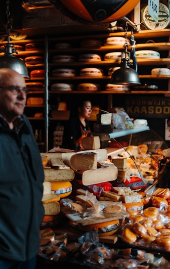 A busy cheese shop displaying a variety of cheeses stacked and arranged on a counter. A person is seen working behind the counter, and another individual is in the foreground. The shop is lit by hanging lamps, and shelves filled with wheels of cheese line the walls.