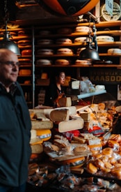 A busy cheese shop displaying a variety of cheeses stacked and arranged on a counter. A person is seen working behind the counter, and another individual is in the foreground. The shop is lit by hanging lamps, and shelves filled with wheels of cheese line the walls.