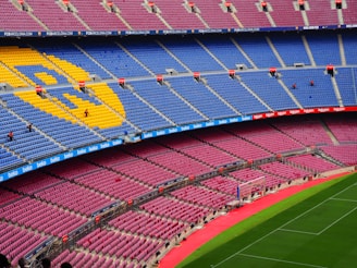 A large, empty football stadium with rows of colorful seats, predominantly blue and red, featuring a large emblem made of yellow seats. The grass pitch is vibrant green, and there are advertising banners along the stands.