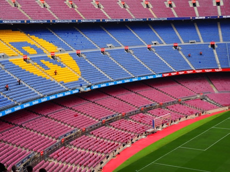 A large, empty football stadium with rows of colorful seats, predominantly blue and red, featuring a large emblem made of yellow seats. The grass pitch is vibrant green, and there are advertising banners along the stands.