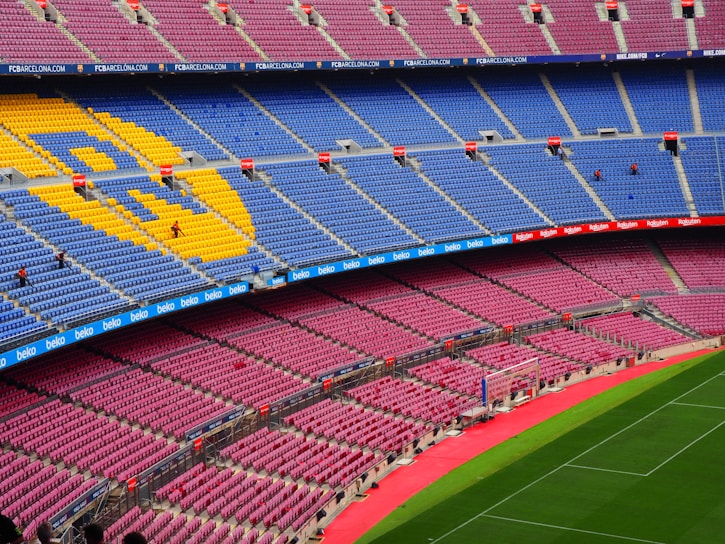 A large, empty football stadium with rows of colorful seats, predominantly blue and red, featuring a large emblem made of yellow seats. The grass pitch is vibrant green, and there are advertising banners along the stands.
