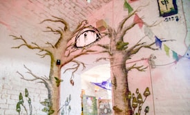 A room with two large mural trees on a white brick wall, branches extending towards the ceiling. An eye symbol with dangling strings is positioned between the tree trunks. Colorful prayer flags hang from the ceiling, and other abstract patterns and shapes decorate the space.
