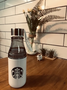 A white water bottle with a black logo on a wooden table, accompanied by a decorative plant in the background. The scene is framed by a white tiled wall, contributing to a clean and modern aesthetic.