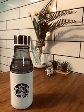 A white water bottle with a black logo on a wooden table, accompanied by a decorative plant in the background. The scene is framed by a white tiled wall, contributing to a clean and modern aesthetic.
