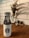 A white water bottle with a black logo on a wooden table, accompanied by a decorative plant in the background. The scene is framed by a white tiled wall, contributing to a clean and modern aesthetic.