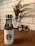 A white water bottle with a black logo on a wooden table, accompanied by a decorative plant in the background. The scene is framed by a white tiled wall, contributing to a clean and modern aesthetic.