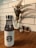 A white water bottle with a black logo on a wooden table, accompanied by a decorative plant in the background. The scene is framed by a white tiled wall, contributing to a clean and modern aesthetic.