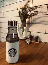 A white water bottle with a black logo on a wooden table, accompanied by a decorative plant in the background. The scene is framed by a white tiled wall, contributing to a clean and modern aesthetic.