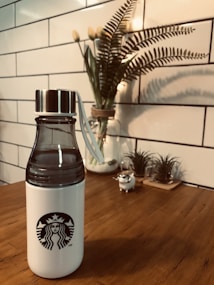 A white water bottle with a black logo on a wooden table, accompanied by a decorative plant in the background. The scene is framed by a white tiled wall, contributing to a clean and modern aesthetic.