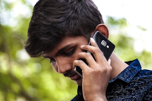 A young man is holding a smartphone to his ear, appearing to be engaged in a phone call. He has short, dark hair and is wearing a dark-colored, textured shirt. The background is blurred but shows green foliage, indicating an outdoor setting.
