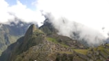 An ancient city with stone structures and terraces set on a steep mountain ridge, partially covered by clouds. Lush green mountains surround the area, and patches of grass are visible among the ruins. A few people are scattered across the site, exploring the historical location.