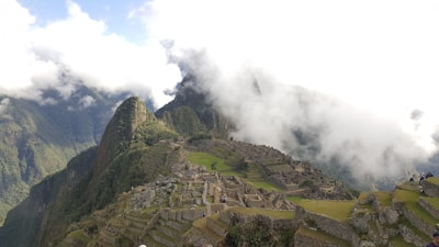 An ancient city with stone structures and terraces set on a steep mountain ridge, partially covered by clouds. Lush green mountains surround the area, and patches of grass are visible among the ruins. A few people are scattered across the site, exploring the historical location.