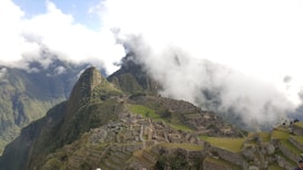 An ancient city with stone structures and terraces set on a steep mountain ridge, partially covered by clouds. Lush green mountains surround the area, and patches of grass are visible among the ruins. A few people are scattered across the site, exploring the historical location.