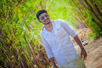 A person is walking along a path surrounded by lush greenery. They are wearing a white shirt and sunglasses, smiling warmly. The sunlight filters through the trees, creating a vibrant and lively atmosphere.