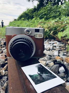 A vintage-style instant camera is placed on a rusty rail among rocks and greenery. In front of it lies a developed photo showing a person standing on railway tracks, surrounded by nature. The background features lush green foliage and a cloudy sky.