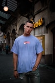 A young man stands in an urban street setting, wearing a blue T-shirt and jeans. Neon signage decorates the background, with architectural elements like arches and ornate details visible on the building. The man has a short haircut and is accessorized with bracelets and a necklace.
